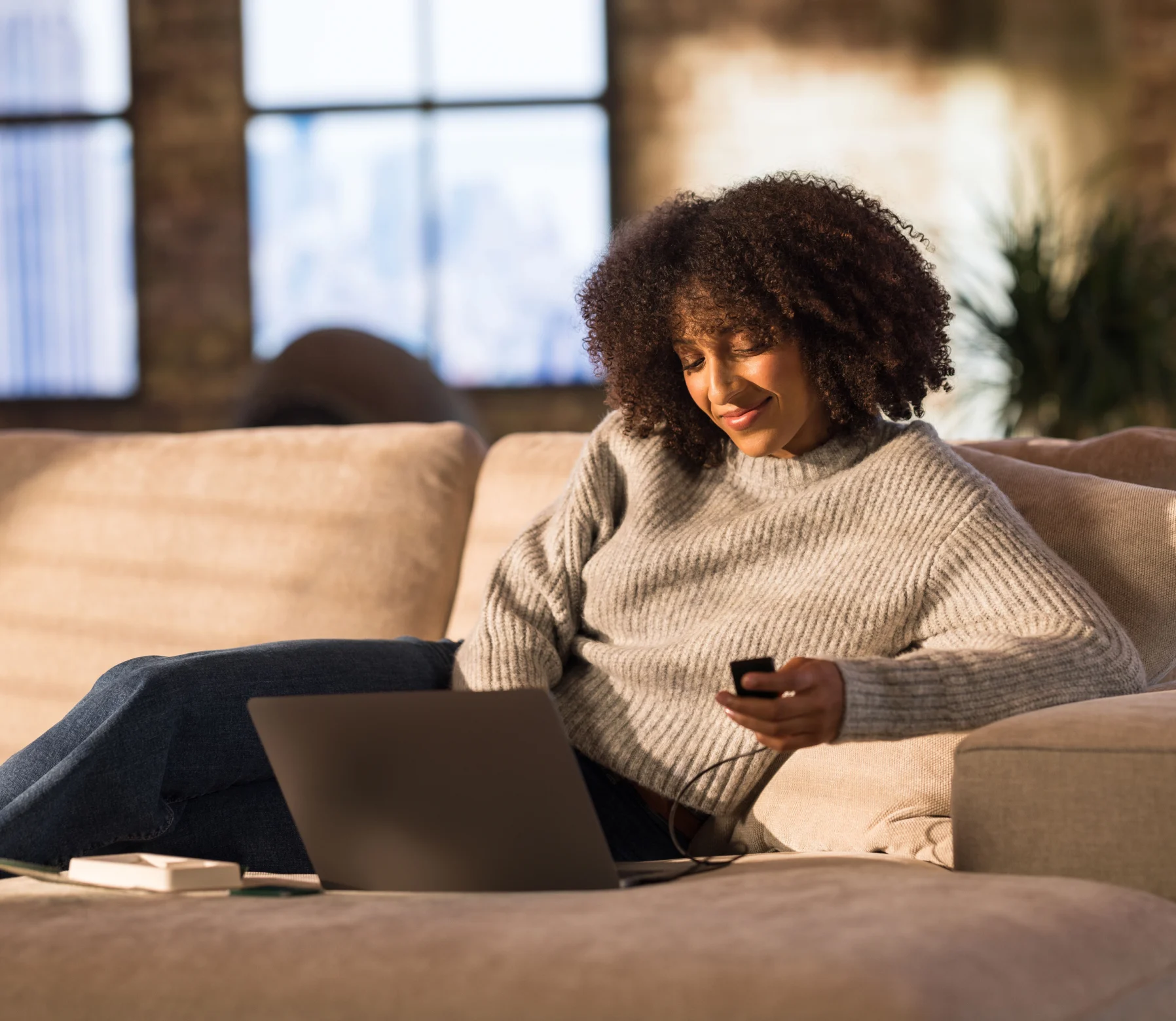 A woman in a cream sweater on a couch with a laptop and phone in a warm, sunlit room.