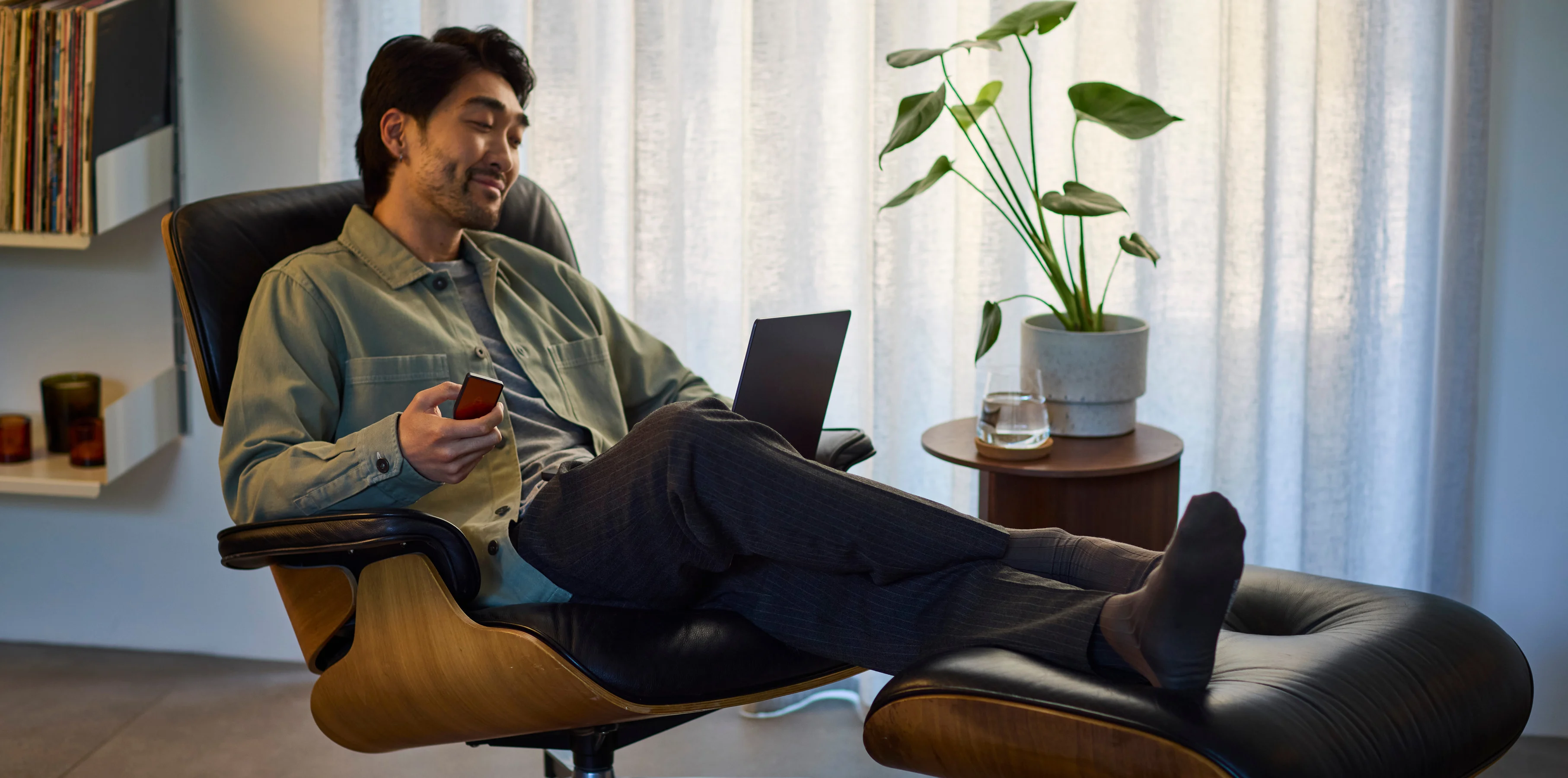 A man reclining in a lounge chair with his phone in a plant-filled, sunlit room.